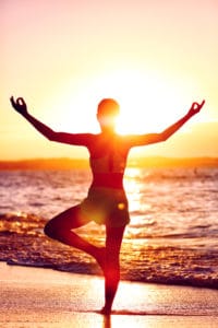 wellness of mind yoga woman standing on one leg doing tree pose with open raised arms in sunset flare in front of the ocean on beach mindfulness and meditation concept